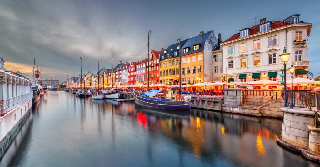 Kopenhag Nyhavn: renkli tarihî cepheler, kanalda ahşap yelkenliler ve sokak lambalarının su üzerindeki yansıması, alacakaranlık.