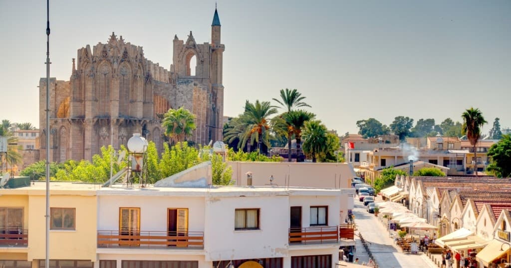 Gotik taş cepheli tarihî cami ve çevredeki beyaz sıvalı binalar, palmiyeler ve açık gökyüzü.