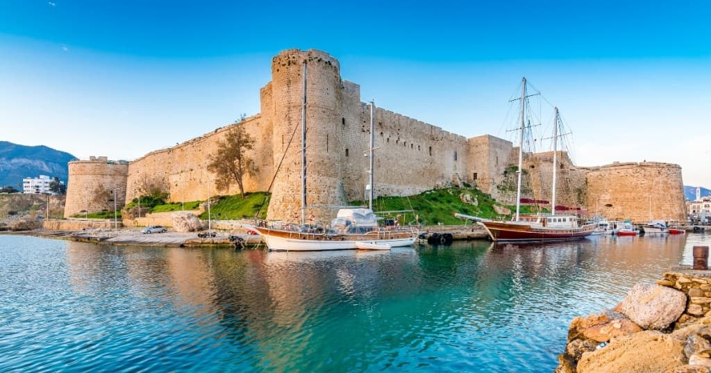 Girne limanı: turkuaz suda ahşap yelkenliler ve kıyıdaki taş Girne Kalesi surları.