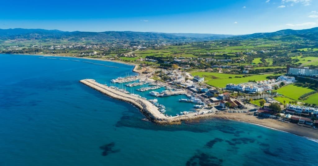 Kıyı marina: kırık dalga koruması içinde beyaz yelkenliler, yeşil sahil ve uzakta dağ silueti.